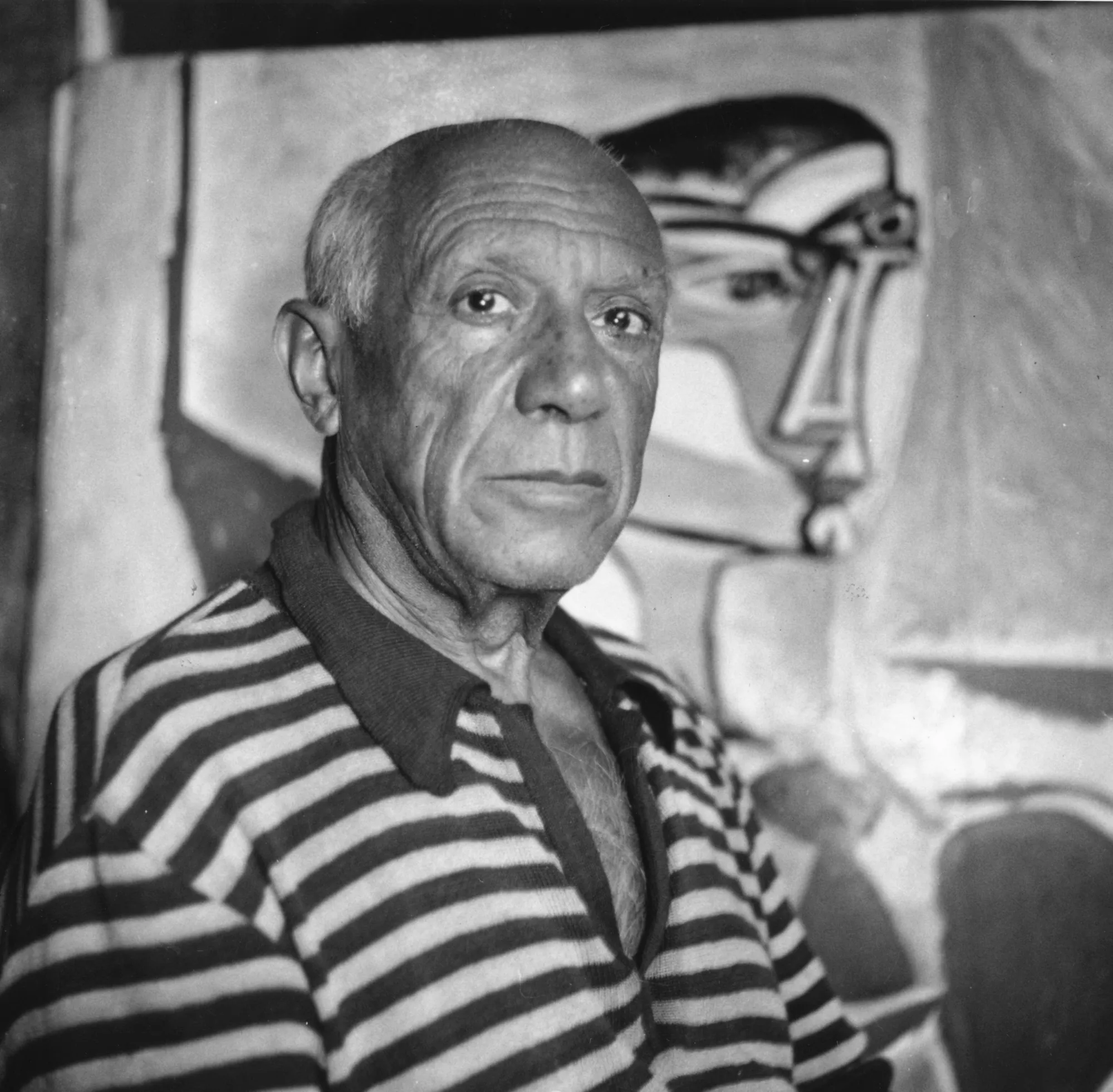 An older man in a striped shirt poses in a black-and-white photo, from the chest up, in front of a painting of an abstracted figure.