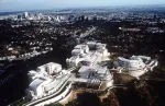 A sprawling modernist structure—all white—sits atop a hit in a wooded area. A city looms in the distance.