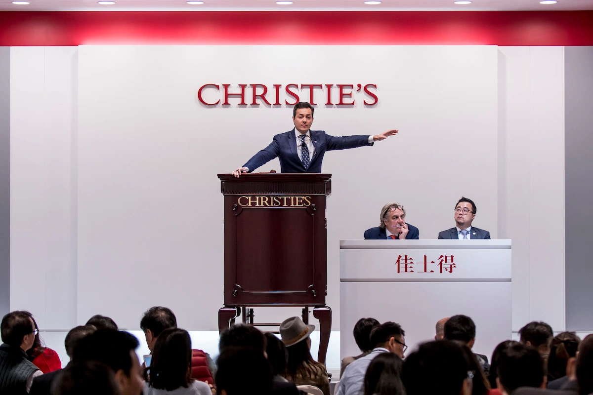 Salesroom with an auctioneer at a podium raising his hand over a crowd.