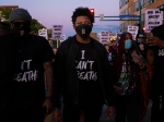 A Black man stands in the center of a color photograph. He wears a face mask and a T-shirt that says "I can't breathe."