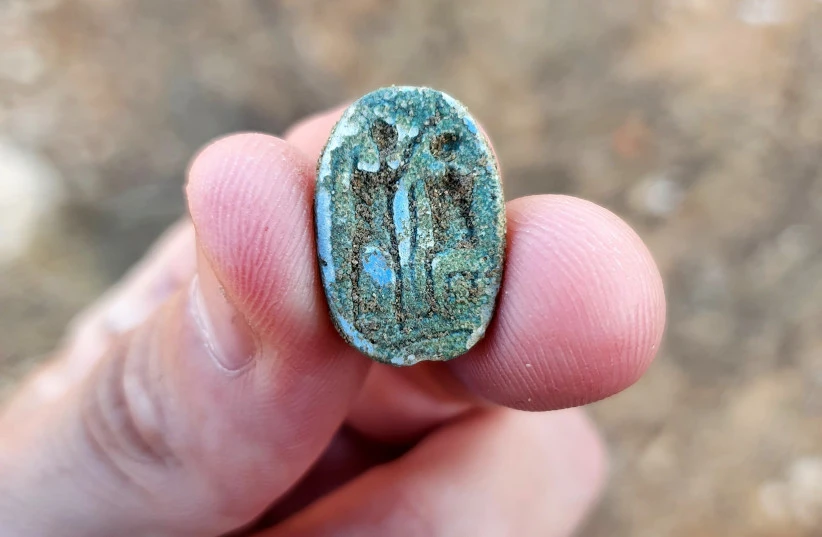 Fingers hold a tiny scarab on which is carved a seated figure on the right and a standing figure with a raised arm on the left.