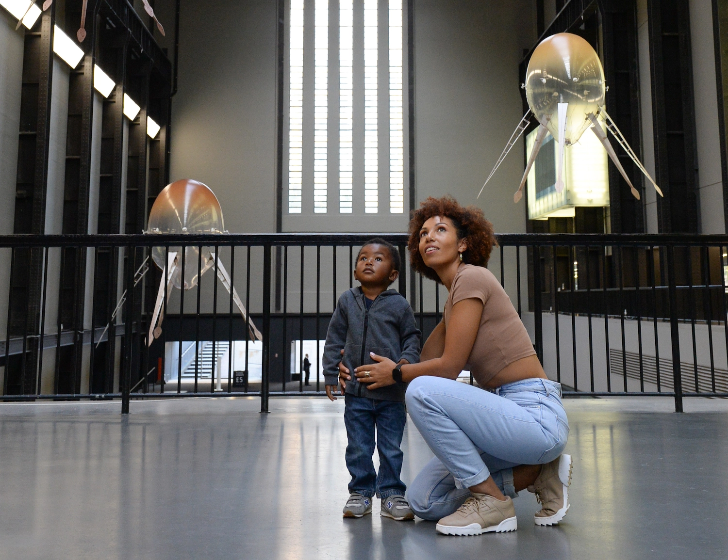 A black woman and a young boy look up at two floating ovoid sculptures.