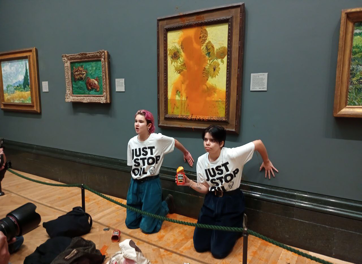 Two protesters, both wearing shirts reading 'JUST STOP OIL,' beneath a painting of sunflowers that has been splashed with soup. They kneel beneath, with their hands stuck to the gallery wall. A group of cameramen photograph them.