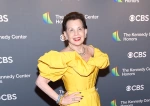 A woman poses in a formal yellow dress, arms akimbo, before a background with the words "CBS" and "The Kennedy Center Honors" repeating behind her.