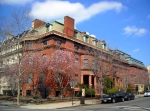 A stately building—dark red in color—with a mansard roof stands on the corner of a street. Nearby trees are bare, save for pink flowers on a few. It is a clear day.