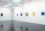 Six modestly sized paintings—geometric abstractions, variously black, green, yellow, and blue—hang on two white walls in a gallery.