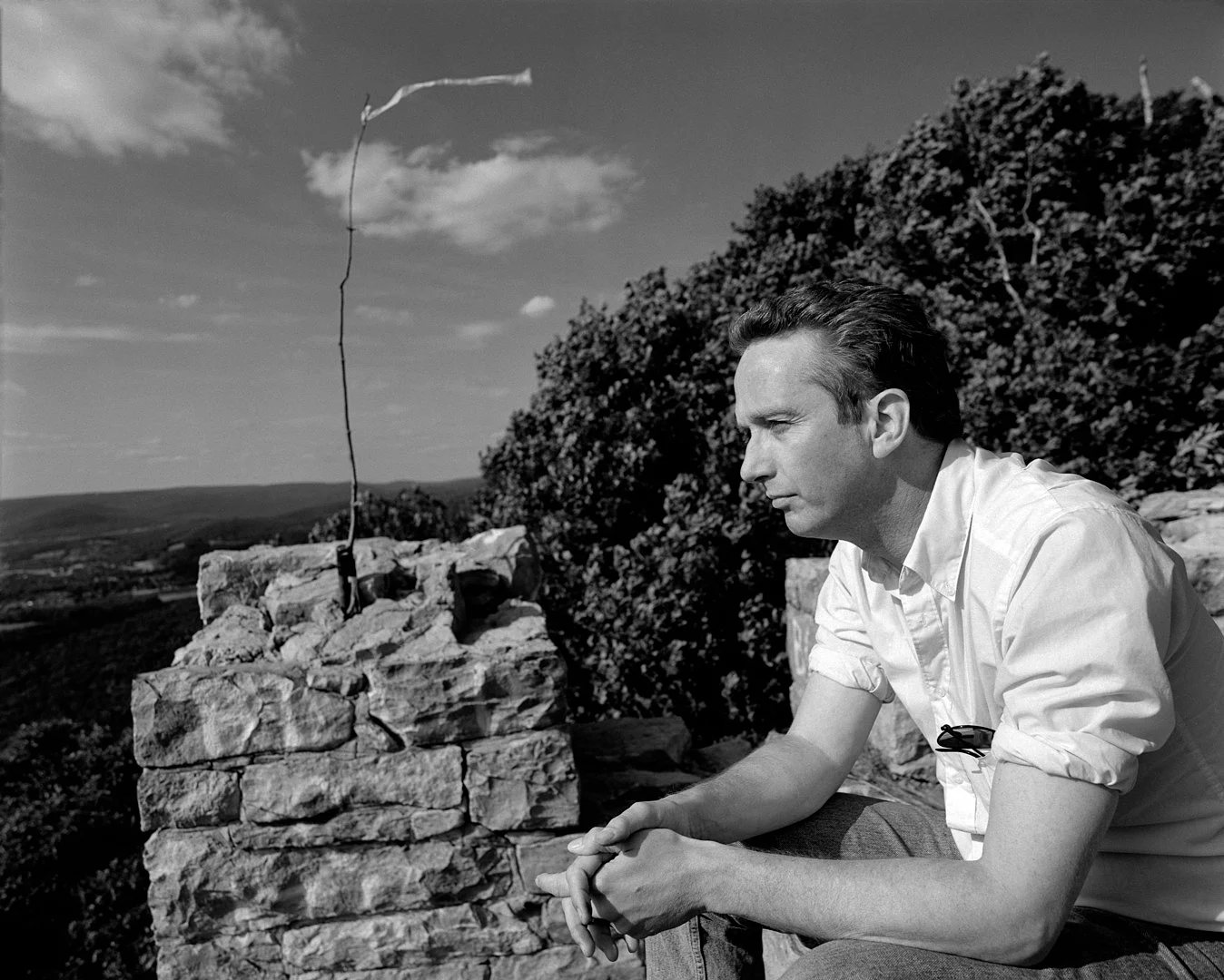 A middle-aged man is seen from the side in a black-and-white photograph. He sits atop something tall, hands folded, as he looks into the distance.
