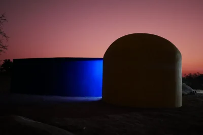 A structure that is almost completely dark seen at dusk, with a pinkish-orange sky. The structure is a yellow dome, silo shape at right that extends to a blue wall at left. 