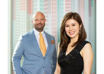 A white man and an Asian woman pose for a photo. He wears a light blue suit with a yellow tie and she wears a black dress. They stand in front of a colorful background. 