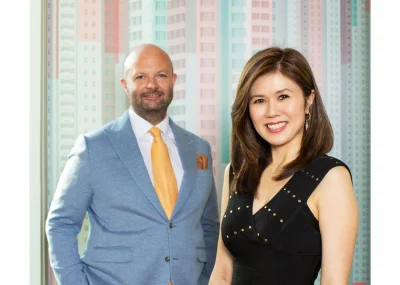 A white man and an Asian woman pose for a photo. He wears a light blue suit with a yellow tie and she wears a black dress. They stand in front of a colorful background.