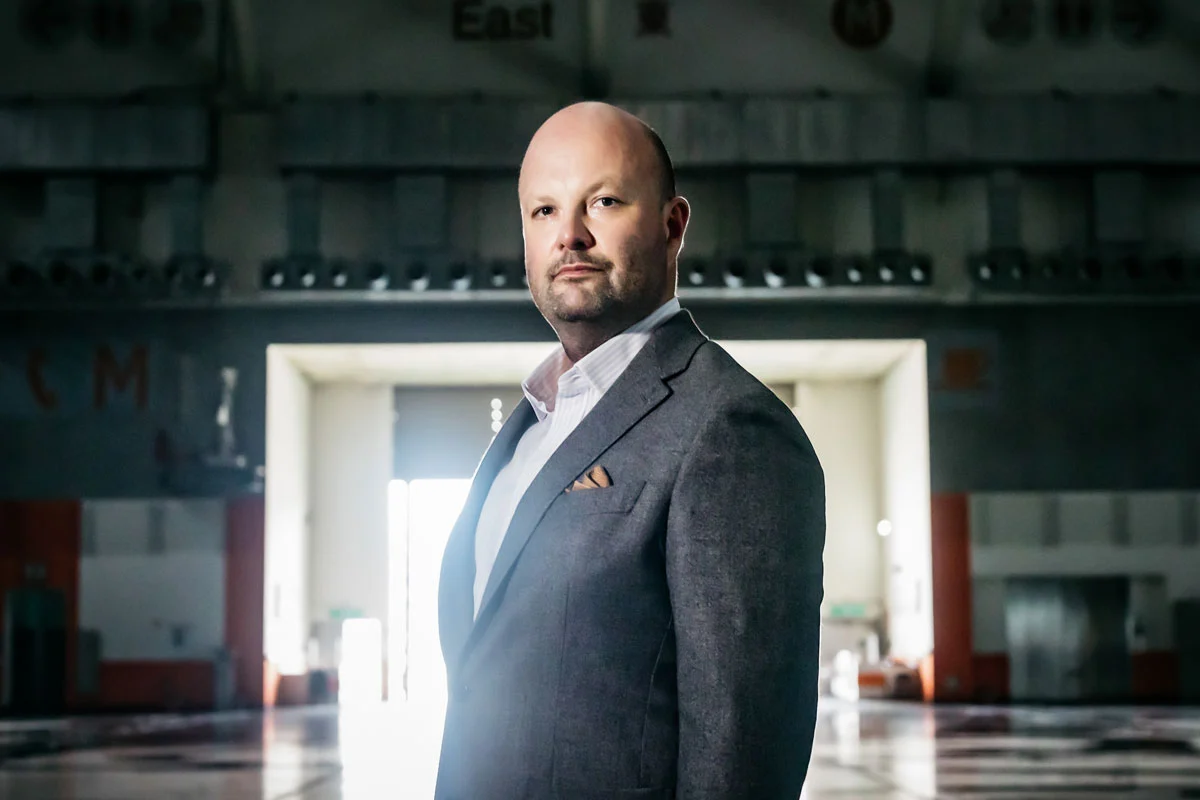 Portrait of a white man who is bald in a gray suit. He stands in the center of a raw convention center space.