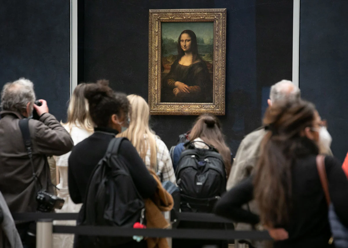 Visitors with their backs to the camera look at the Mona Lisa painting, which hangs on a blue-wall.