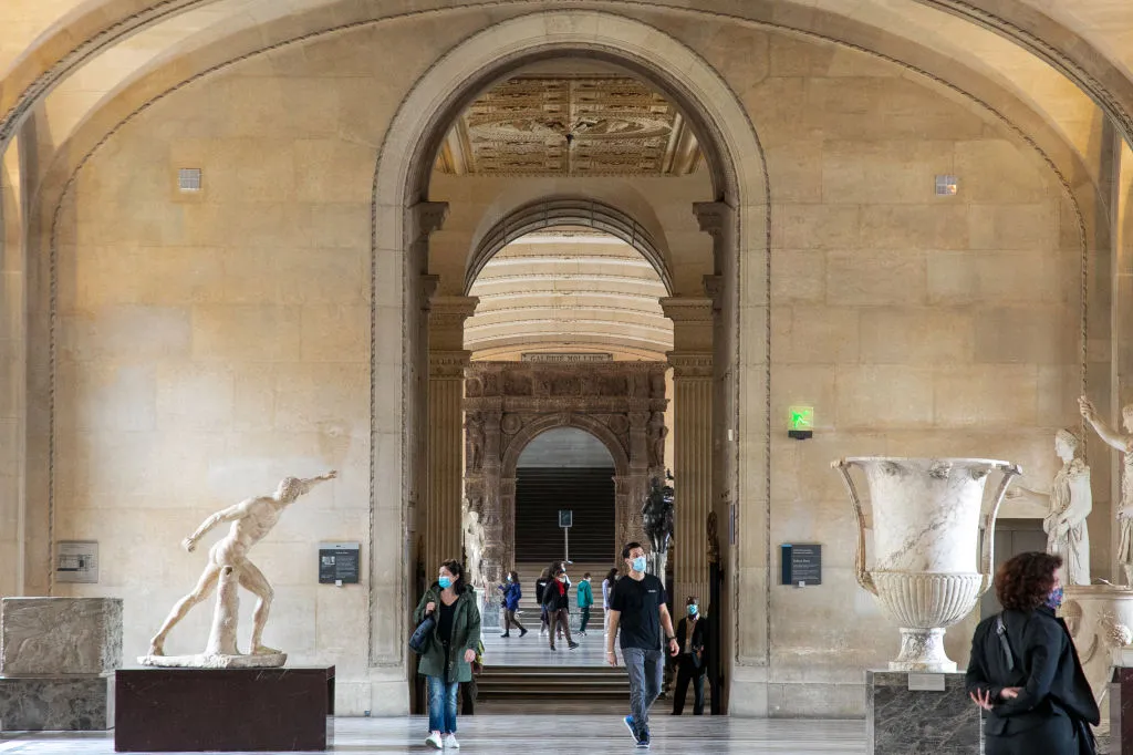 A few people wearing masks walk through a grand series of halls. A few majestic ancient Greek-looking sculptures and artifacts stand on plinths near them.