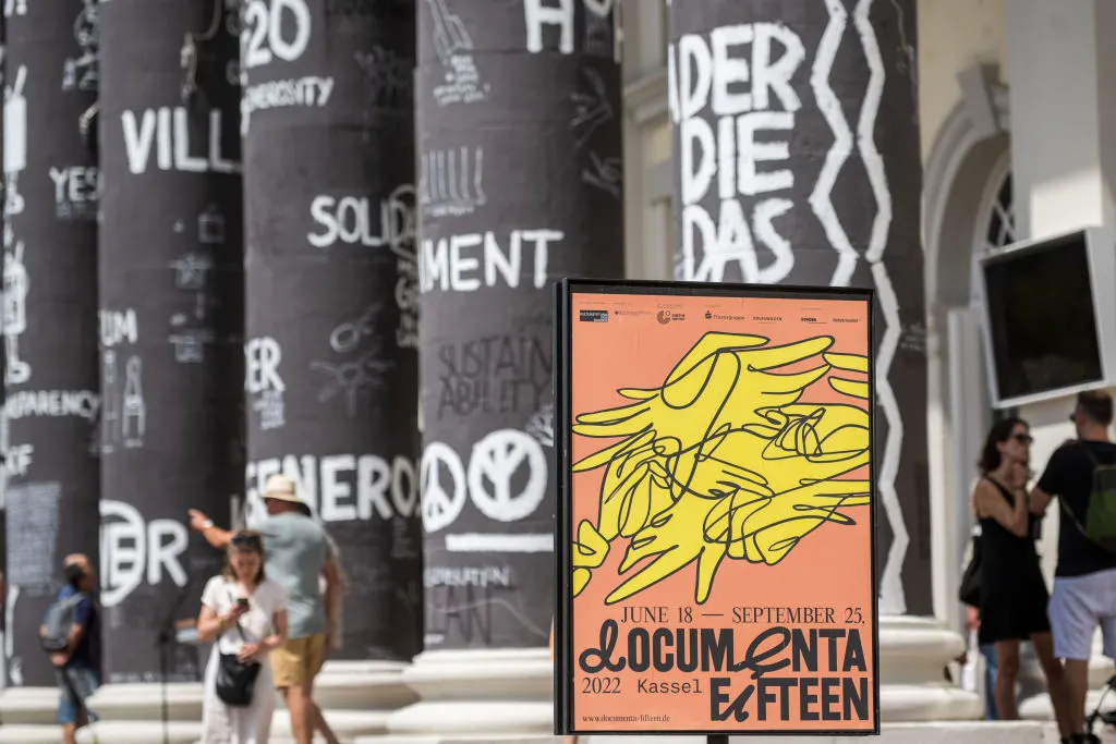 A poster for Documenta 15 appears in a color photograph in front of a number of columns covered in black with white writing.