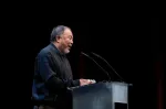 A man stands on stage in a black shirt and speaks at a podium with two microphones.