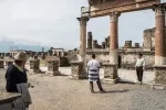 A few people stand amid Roman ruins: partially reconstructed columns, and others that are nearly destroyed.