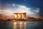 Exterior view of a building seen from across a harbor. There are several low-lying buildings in front of three soaring towers that gleam yellow in the sunset. The towers are connected by one long outdoor balcony that sits atop them.