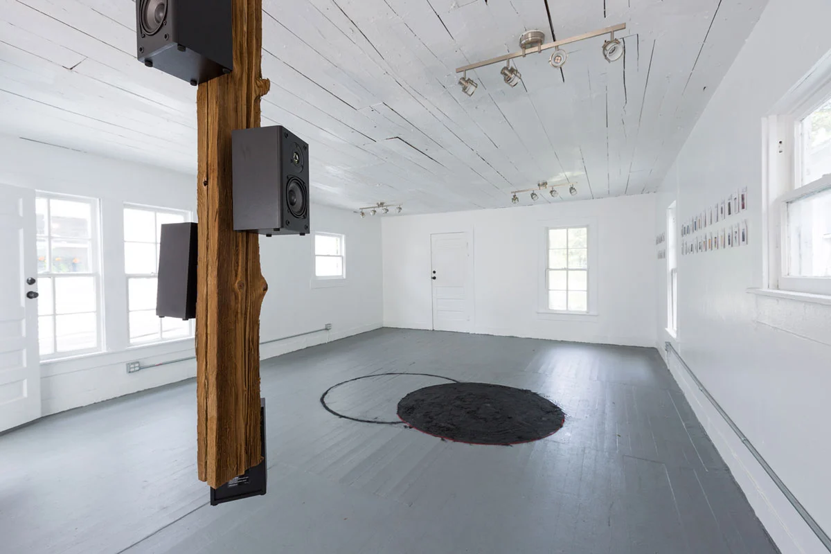Installation view of a white-cube gallery showing a wooden beam to which three speakers are attached. At the center of the gallery is a pile of black charcoal in a circle and to its left is an outline of a circle also in black.