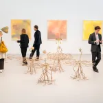 Sculptures of tree roots take over an art fair booth, with two abstract paintings visible in the background. People walk throughout the booth.