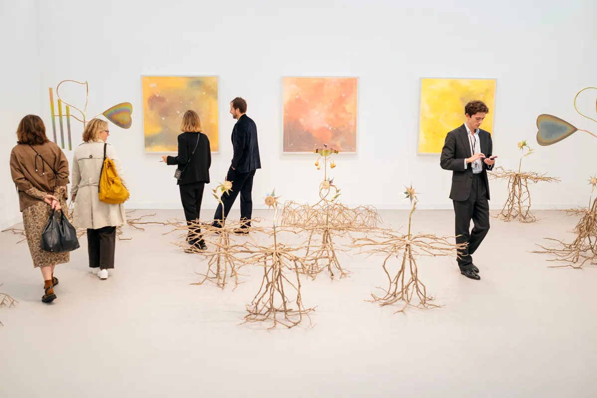 Sculptures of tree roots take over an art fair booth, with two abstract paintings visible in the background. People walk throughout the booth.