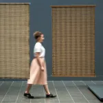 A woman in a white shirt and pink skirt walks past two large brown weavings hanging vertically on a blue wall