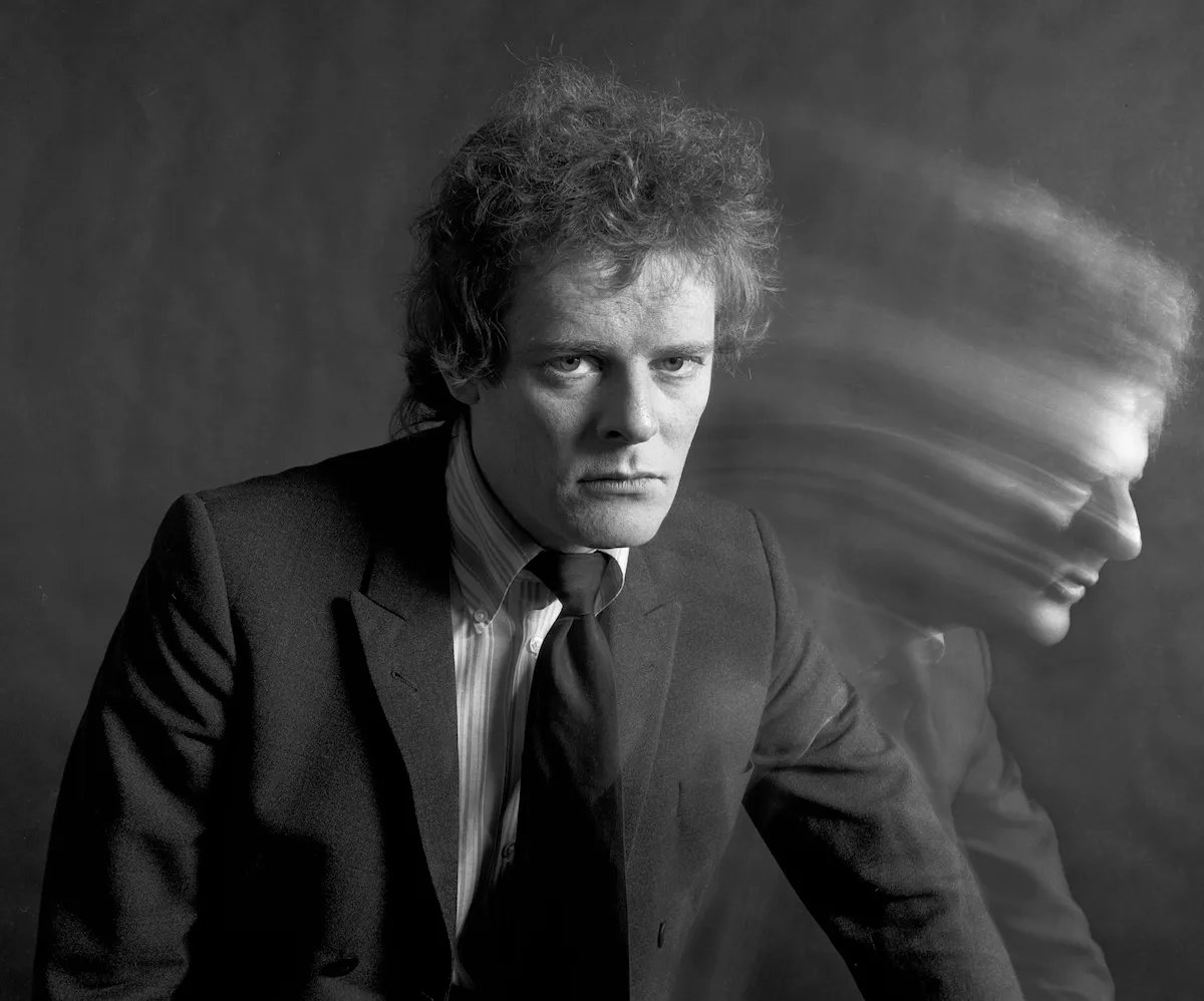 A white man in a suit with a blurred image of himself next to him.