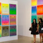 Two women walking amid gridded paintings of smiling faces.