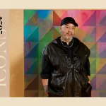 A man in a black vest over a black shirt in a black hat standing in front of a multi-colored abstract painting in the works.