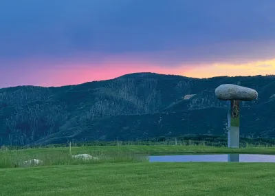 A metal plinth holding a very large rock that is installed near a lake on a ranch with mountains in the background.