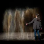 An ephemeral shape that looks like mist meets gold and silver horse hair is suspended in a dark gallery. A long-haired person stands with their back to the camera in front of the shape, showing you that it