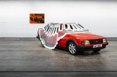 A red Ford Escort draped with a large-scale doily, with a painting in the background.