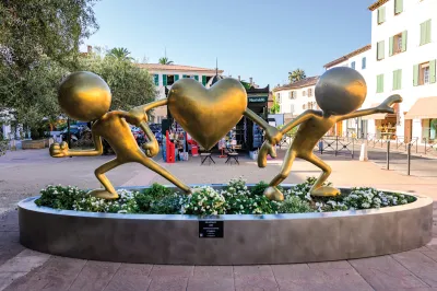 A gold-like sculpture of two stick figures running away from each other with one of their arms each being held by a heart with arms.