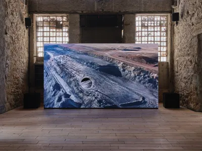 A video projected onto a false-wall screen, showing a desert landscape. It is installed in a rom with rough brick walls.