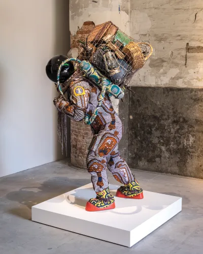 A sculpture showing an astronaut-like figure figure with a net filled with items on his back. His clothing is made of colorful fabrics from Africa.