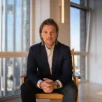 A man in a suit leans forward in a chair as he looks at the camera.