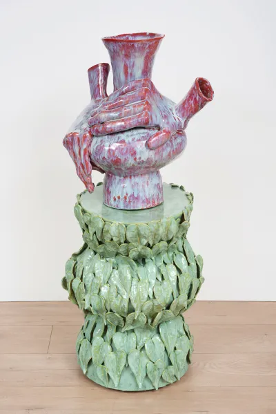 A glazed ceramic sculpture featuring two hands working on a vase in red, light blue, and white; this sits on a base of light-green leaves.