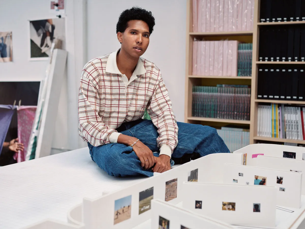 Portrait of Tyler Mitchell, sitting on a table in his studio looking at a maquette for an exhibition.