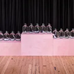 Three award-ceremony-like platforms in pink hold five glass jugs filled with a white ribbon.