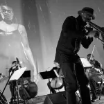 Musicians play in front of a screen that shows a Black woman standing in the rain.