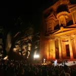 An ancient structure carved into a mountain lit up at night with a crowd in front of it.