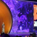 A woman in a dress stands on stage next to a large screen.