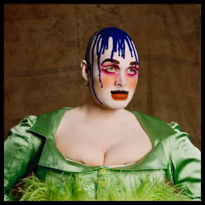 A person with blue goo streaming down over their head posing for the camera. They wear a green dress that just barely conceals their breasts.