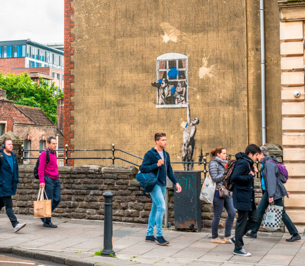 A building with a mural of a naked man hanging out of a window as people walk by.