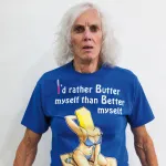 A woman with long white hair with a surprised expression wearing a blue T-shirt that reads 