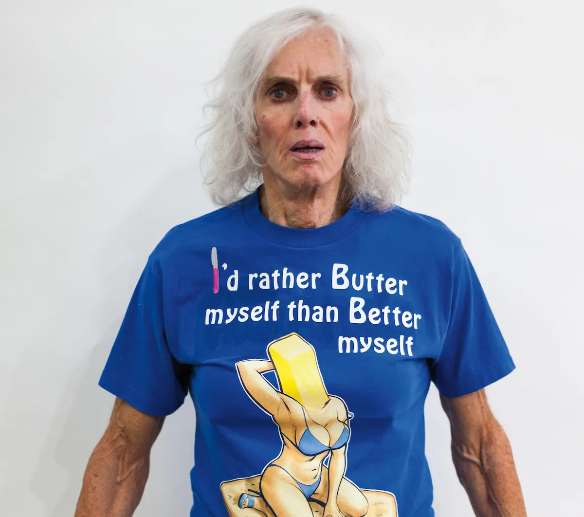 A woman with long white hair with a surprised expression wearing a blue T-shirt that reads