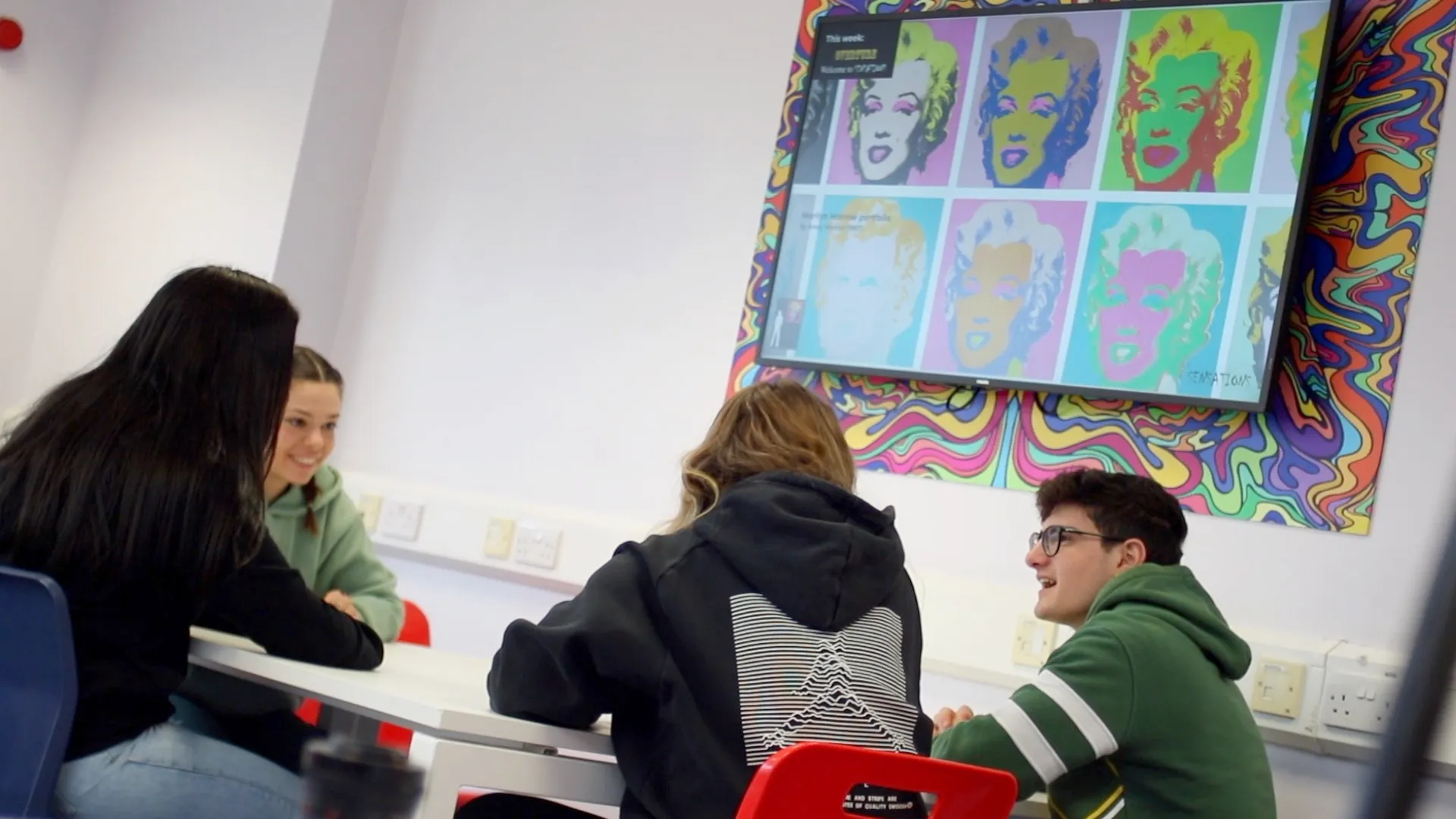 Four students talk beneath a screen showing art.