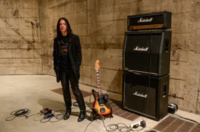 A white woman in black leather pants standing next to a guitar and three stacked amplifiers.