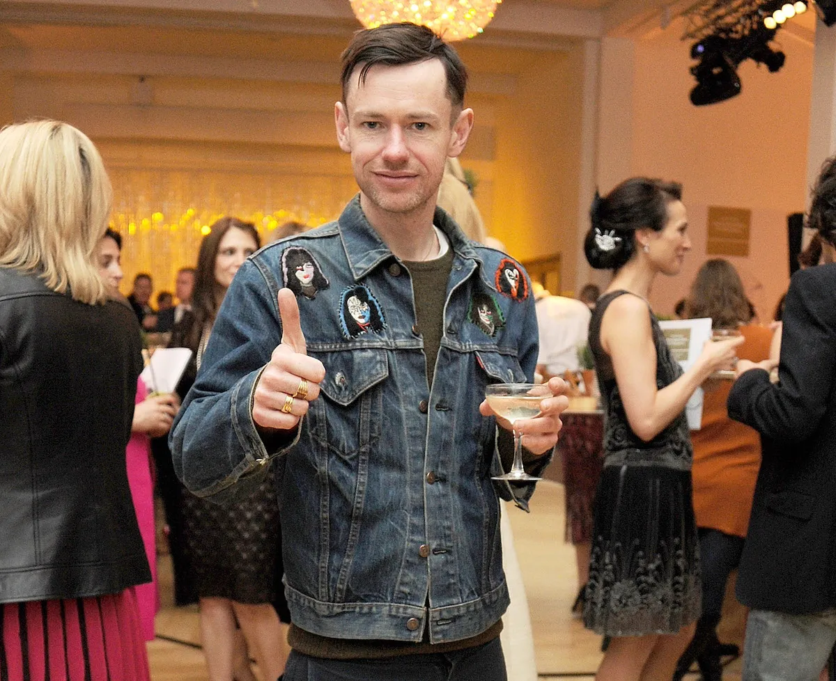 A white man in a denim jacket holding up one thumb with one hand and a martini glass in the other.