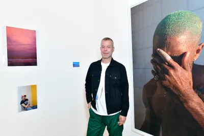 A white man in a black jacket standing beside a large photograph of a Black man with green hair clutching a hand to his face.