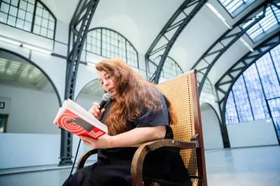 A woman in a wicker chair, leaning over a book and speaking into a microphone.